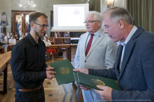 Wręczenie dokumentów w sali obrad, trzech mężczyzn w formalnych strojach.