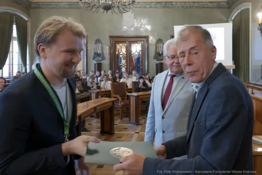 Wręczenie nagrody w sali obrad, uśmiechnięci uczestnicy, dokument w dłoniach.