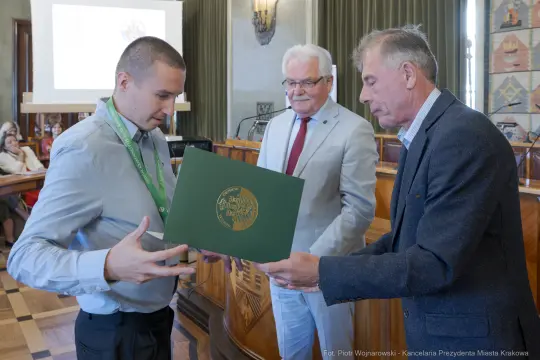 Wręczenie dyplomu w sali obrad, trzech mężczyzn, jeden z medalem, dokument w rękach.