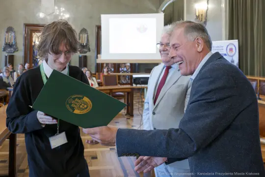 Wręczenie dyplomu podczas uroczystości w sali obrad. Uczestnicy uśmiechnięci.