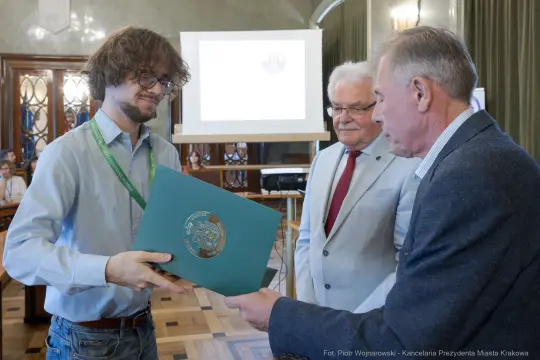 Mężczyzna odbierający dyplom w obecności dwóch innych mężczyzn w sali konferencyjnej.