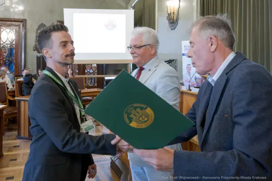 Mężczyzna odbierający dokument w obecności dwóch innych mężczyzn w sali konferencyjnej.