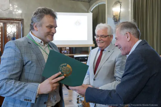 Wręczenie dyplomu w sali konferencyjnej, uśmiechnięci mężczyźni w formalnych strojach.