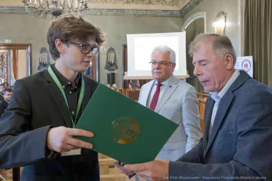 Młody mężczyzna odbiera dyplom od starszego mężczyzny w formalnym otoczeniu.