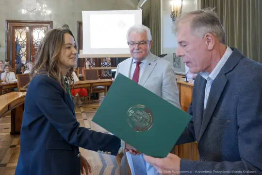 Wręczenie dyplomu w sali obrad, uśmiechnięte osoby, dokument w rękach.