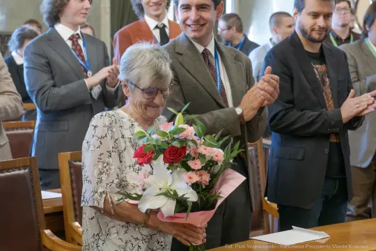 Starsza kobieta trzyma bukiet kwiatów, otoczona przez uśmiechniętych ludzi.