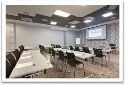Pusty pokój konferencyjny z ustawionymi stołami i krzesłami, ekranem oraz materiałami do pisania.