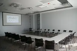 Sala konferencyjna z okrągłym układem stołów i krzeseł, ekranem projekcyjnym.
