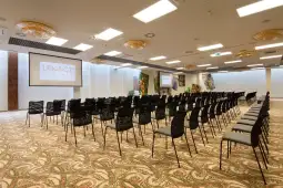 Sala konferencyjna z rzędem czarnych krzeseł i ekranem projekcyjnym.