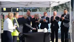 Uroczystość symbolicznego wbicia łopaty na budowie, uczestnicy w garniturach i kaskach.