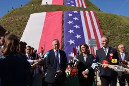 Uroczystość z flagami Polski i USA oraz mówcami przy wzgórzu.