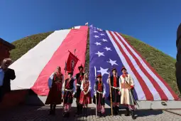 Grupa ludzi w tradycyjnych strojach, trzymających flagi Polski i USA, przed wzgórzem.