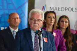 Mężczyzna w garniturze przemawia do mikrofonu, w tle grupka osób.