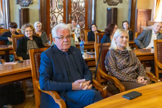 Grupa osób siedzących w sali, uczestniczących w spotkaniu lub konferencji.