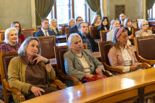 Grupa osób siedzących w sali, skupionych na wydarzeniu.