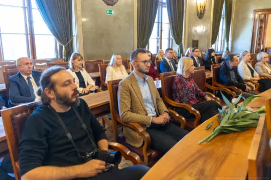 Grupa ludzi siedzących w sali, niektórzy z kamerami, w trakcie wydarzenia formalnego.
