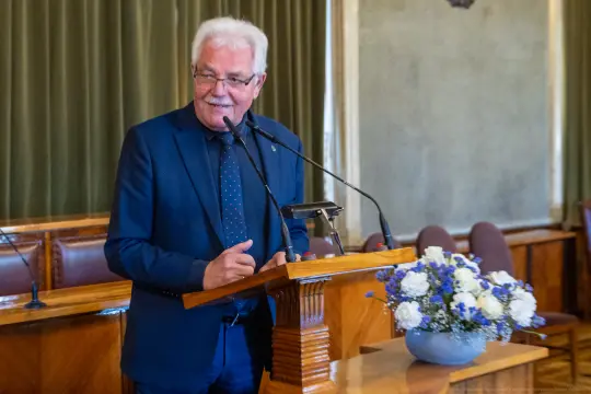 Mężczyzna w garniturze przemawia przy mównicy, obok bukiet kwiatów.