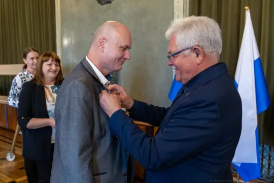 Mężczyzna otrzymuje odznaczenie, w tle widoczna grupa osób oraz flagi.