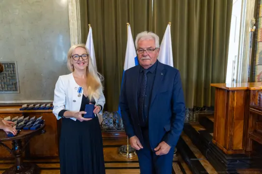 Wręczenie odznaczeń w eleganckim wnętrzu, dwie osoby w formalnych strojach.