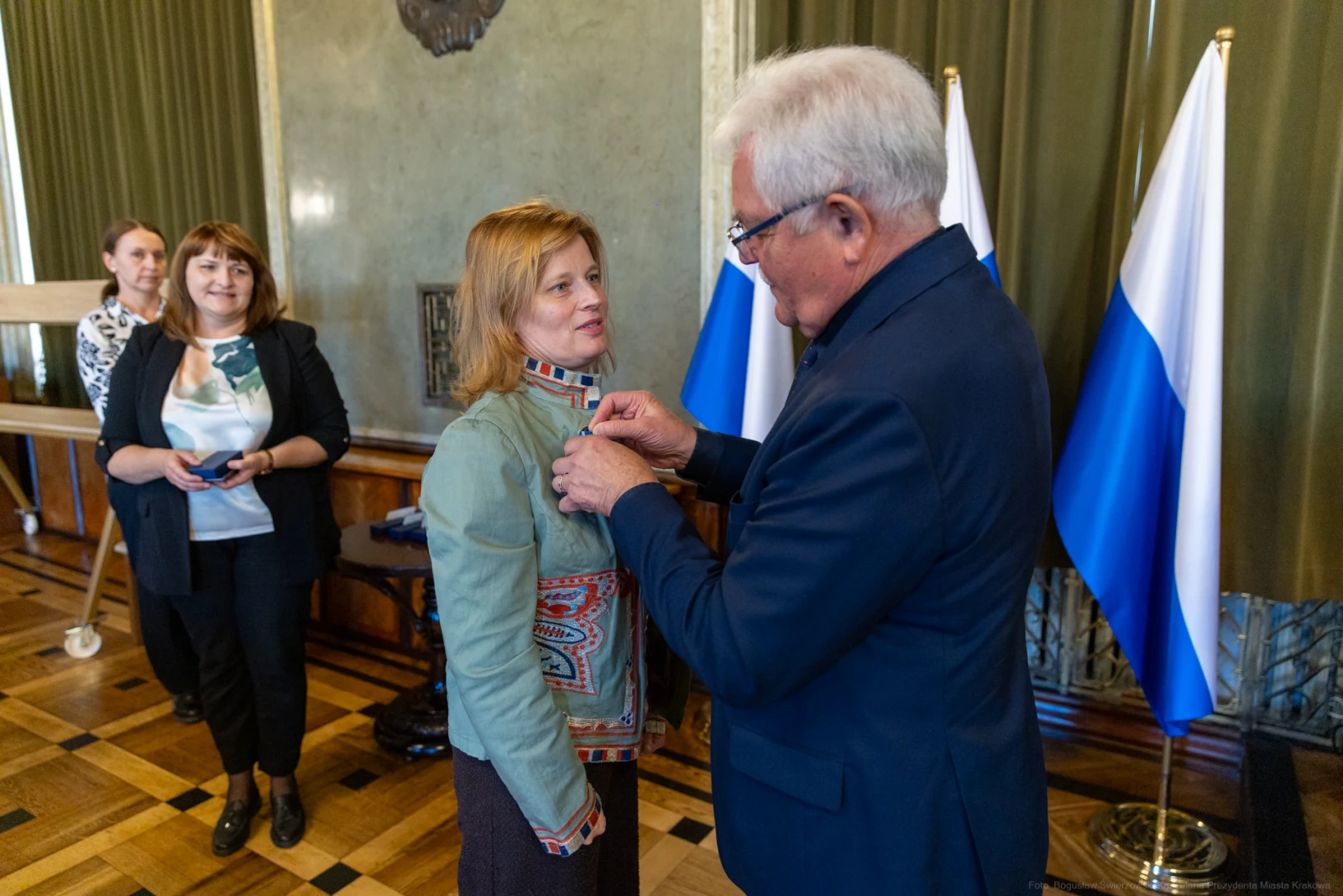 Wręczenie odznaczenia kobiecie przez mężczyznę w formalnym otoczeniu z flagami.  Autor: Bogusław Świerzowski