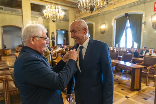 Dwaj mężczyźni w garniturach uśmiechają się i rozmawiają w eleganckim wnętrzu sali.