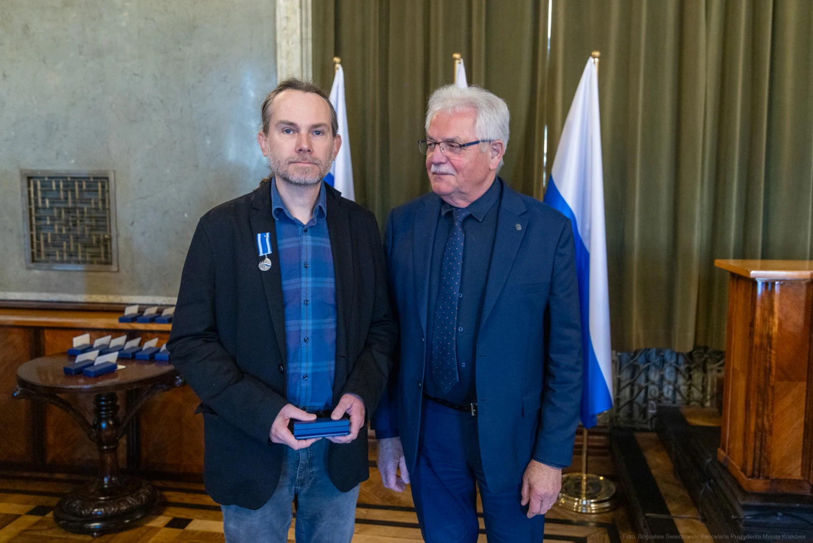 Mężczyzna z odznaczeniem stoi obok drugiego mężczyzny w eleganckim stroju, w tle flagi.  Autor: Bogusław Świerzowski