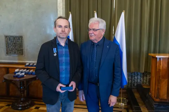 Mężczyzna z odznaczeniem stoi obok drugiego mężczyzny w eleganckim stroju, w tle flagi.