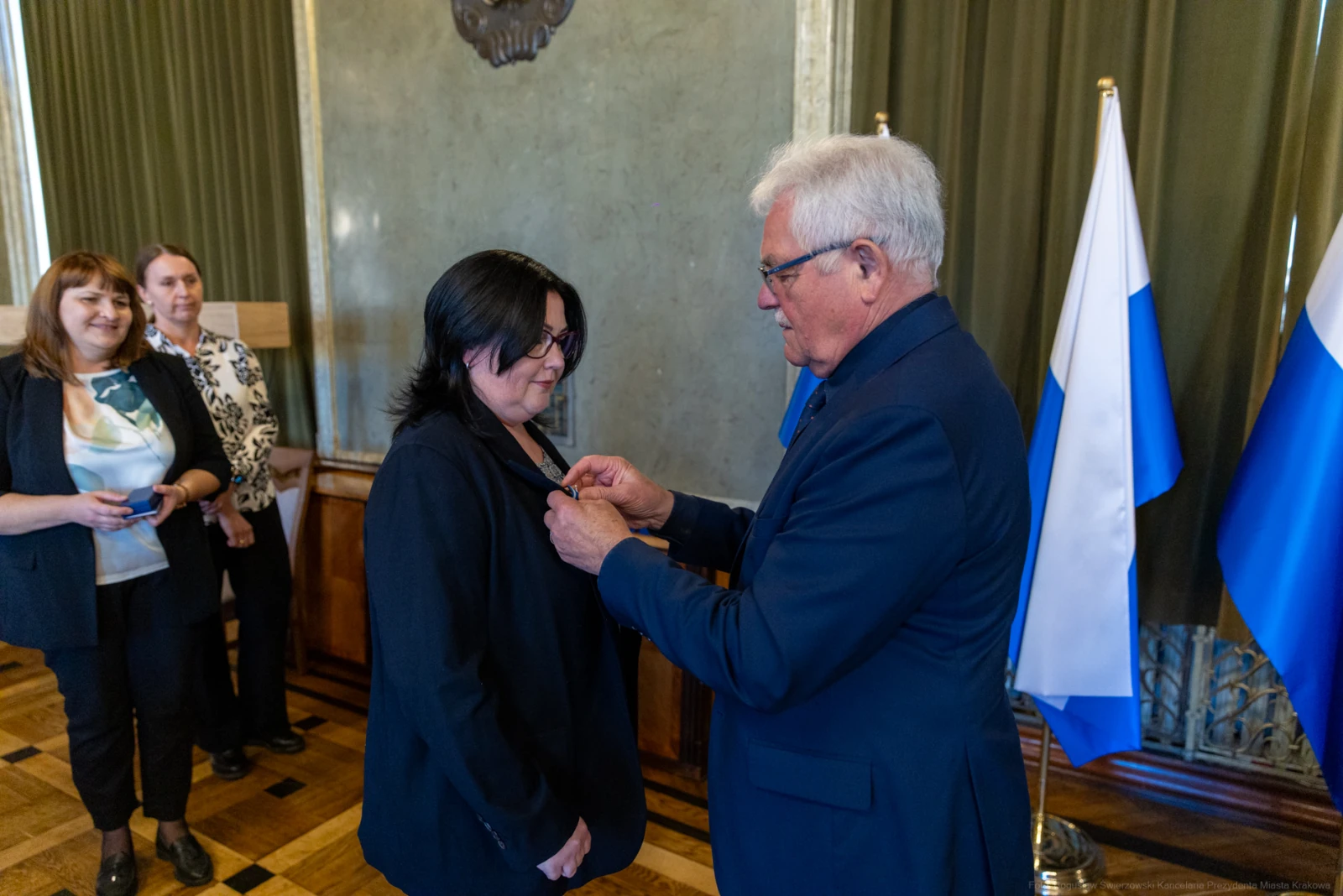 Wręczenie odznaczenia przez mężczyznę kobiecie w obecności innych osób. Flagi w tle.  Autor: Bogusław Świerzowski