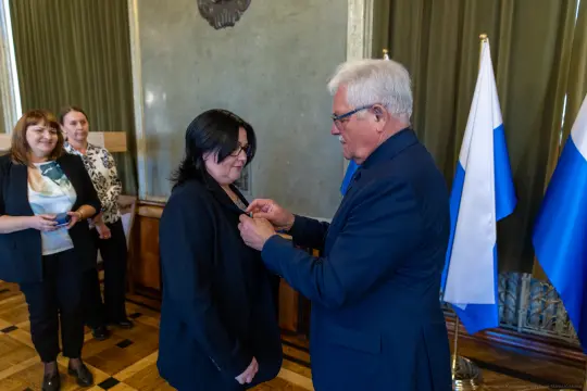 Wręczenie odznaczenia przez mężczyznę kobiecie w obecności innych osób. Flagi w tle.