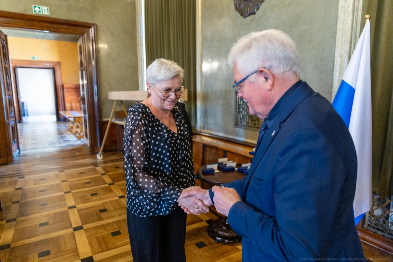 Wręczenie odznaczenia w eleganckim wnętrzu z flagą w tle.  Autor: Bogusław Świerzowski