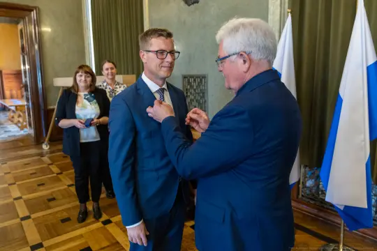 Ceremonia wręczenia odznaczenia mężczyźnie w garniturze, w tle osoby obserwujące wydarzenie.