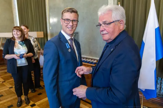 Dwaj mężczyźni w garniturach, jeden z odznaczeniem, w tle kobiety i flagi.