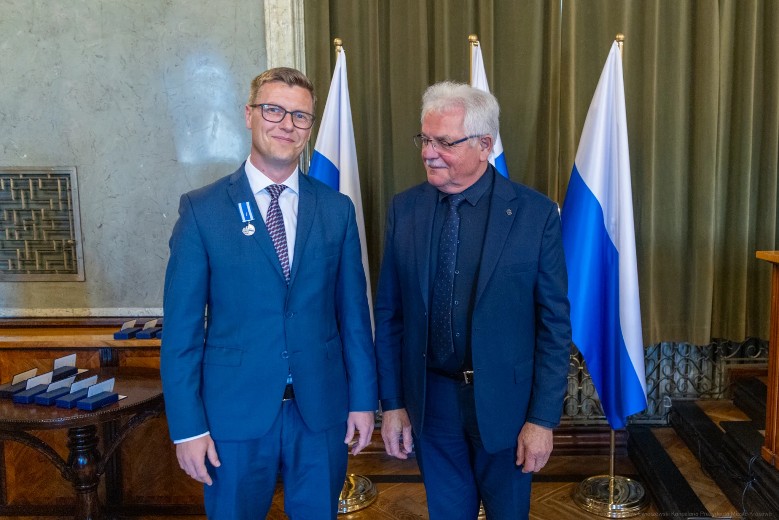 Dwaj mężczyźni w garniturach, jeden z medalem, stoją obok flagi.  Autor: Bogusław Świerzowski