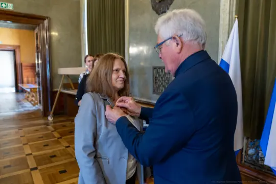 Wręczenie odznaczenia kobiecie przez mężczyznę w eleganckim wnętrzu.