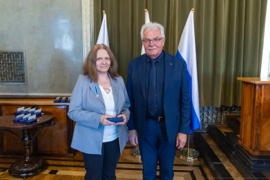 Wręczenie odznaczenia przez mężczyznę kobiecie w eleganckim wnętrzu z flagami.