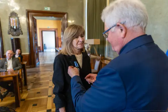 Wręczenie odznaczenia kobiecie przez mężczyznę w eleganckim wnętrzu.