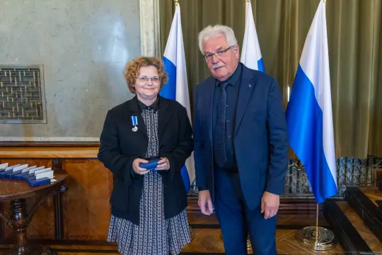 Wręczenie odznaczenia przez mężczyznę kobiecie w eleganckim wnętrzu z flagami.