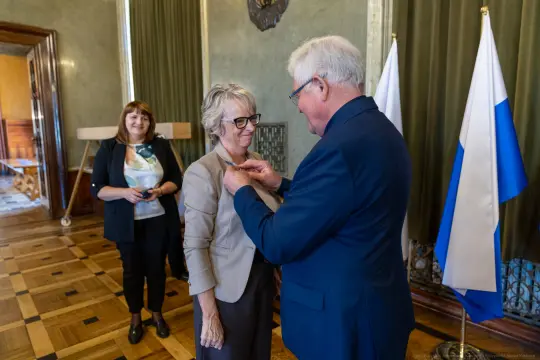 Wręczenie odznaczenia kobiecie przez mężczyznę, w tle flagi i uśmiechnięta osoba.