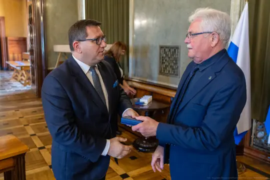 Wręczenie odznaczenia w eleganckim wnętrzu z flagami w tle.