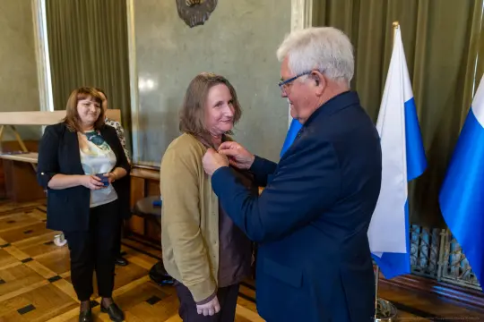 Wręczenie odznaczenia przez mężczyznę kobiecie w sali z flagami.