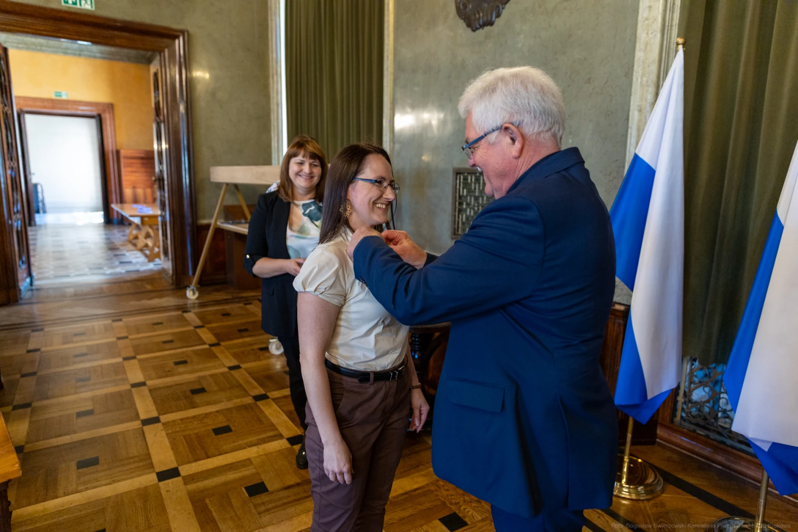 Wręczenie odznaczenia kobiecie przez mężczyznę w eleganckim wnętrzu. W tle widoczna inna osoba.  Autor: Bogusław Świerzowski