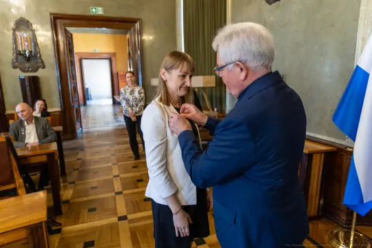 Wręczenie odznaczenia kobiecie przez mężczyznę w formalnym otoczeniu.