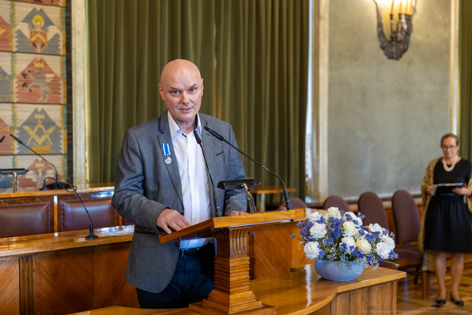 Mężczyzna w garniturze przemawia przy mównicy, obok kwiaty w wazonie.  Autor: Bogusław Świerzowski