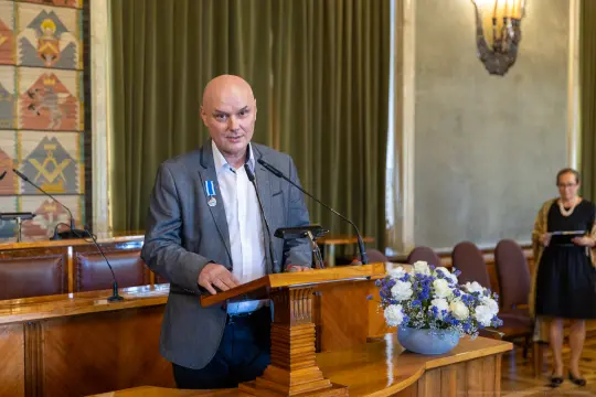 Mężczyzna w garniturze przemawia przy mównicy, obok kwiaty w wazonie.