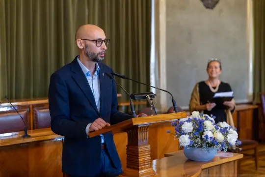 Mężczyzna przemawia z mównicy, w tle kobieta z dokumentami i kwiaty w wazonie.