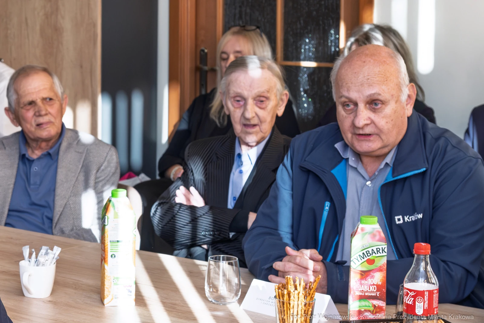 Trzech mężczyzn siedzących przy stole, jeden z nich mówi, w tle widoczni inni uczestnicy spotkania.  Autor: Piotr Wojnarowski
