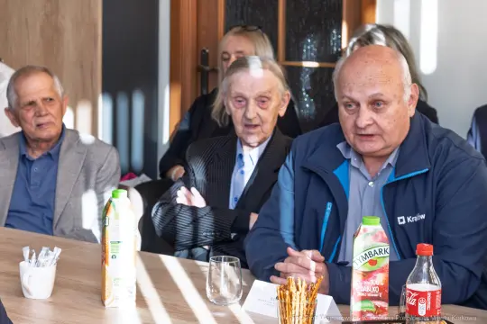 Trzech mężczyzn siedzących przy stole, jeden z nich mówi, w tle widoczni inni uczestnicy spotkania.