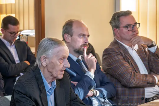 Grupa mężczyzn w trakcie dyskusji, jeden z nich w wieku starszym, pozostali w garniturach.