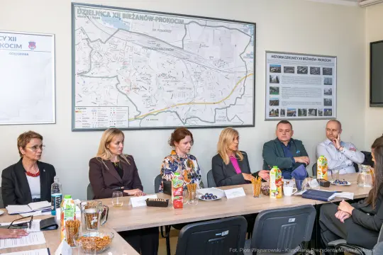 Spotkanie w sali konferencyjnej z mapą dzielnicy i uczestnikami przy stole.