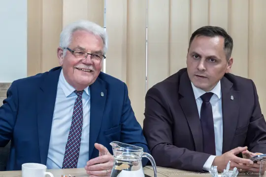 Dwaj mężczyźni w garniturach siedzą przy stole, uśmiechają się i rozmawiają.